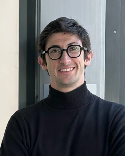 Filippo Saretta, fondatore di Vinary: ragazzo sorridente con occhiali dalla montatura nera e maglione nero a collo alto, a braccia conserte davanti a una parete grigia.